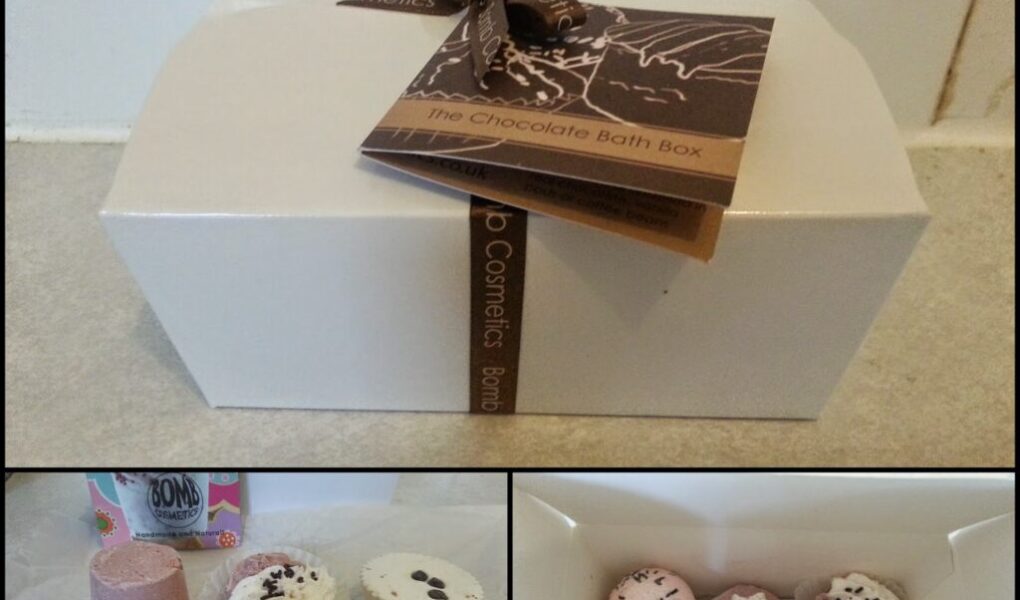 A rectangular white box with a brown ribbon and tag reading “The Chocolate Bath Box.” Below, bath bombs in pink, white, and brown shades, some with decorative toppings, are arranged on a surface and inside an open box.