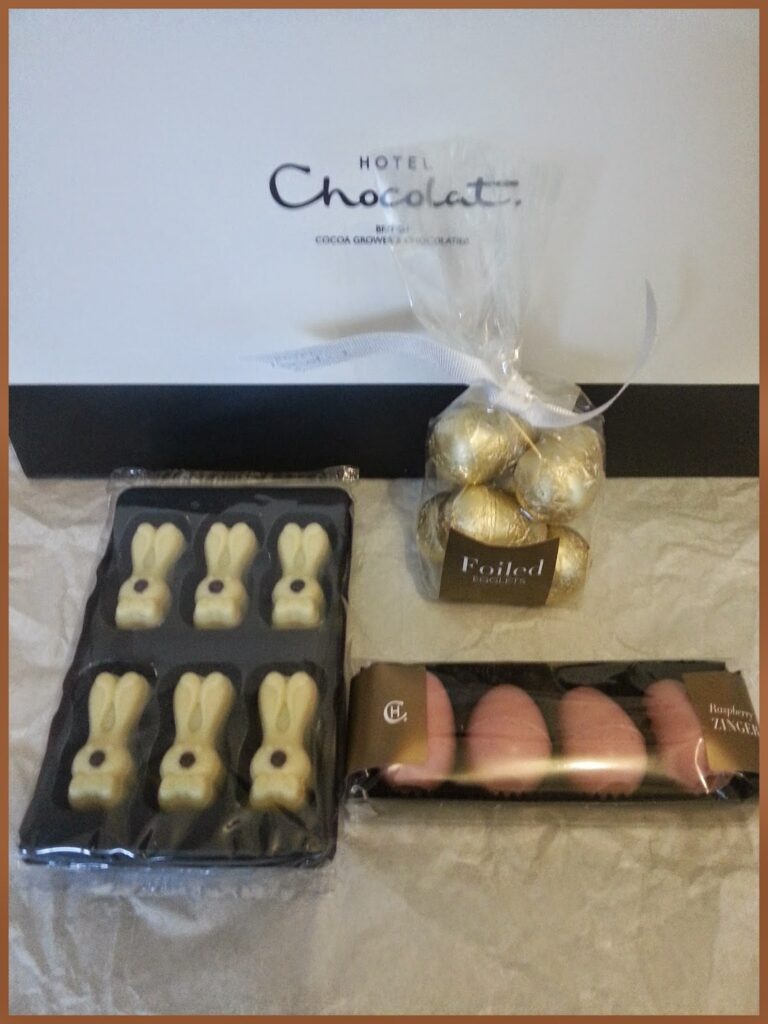 A selection of Hotel Chocolat treats: six white chocolate bunny shapes in a black tray, six gold-foil-wrapped chocolates in a clear bag with a white ribbon, and five pink egg-shaped chocolates in a black tray, all set against a branded box.
