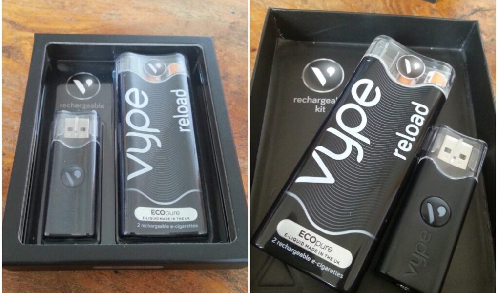 A black box contains a Vype Reload rechargeable e-cigarette kit. Inside are a USB charger and a rectangular black e-cigarette device labelled “vype reload.” Both items have matching branding. The box and its contents rest on a wooden surface.