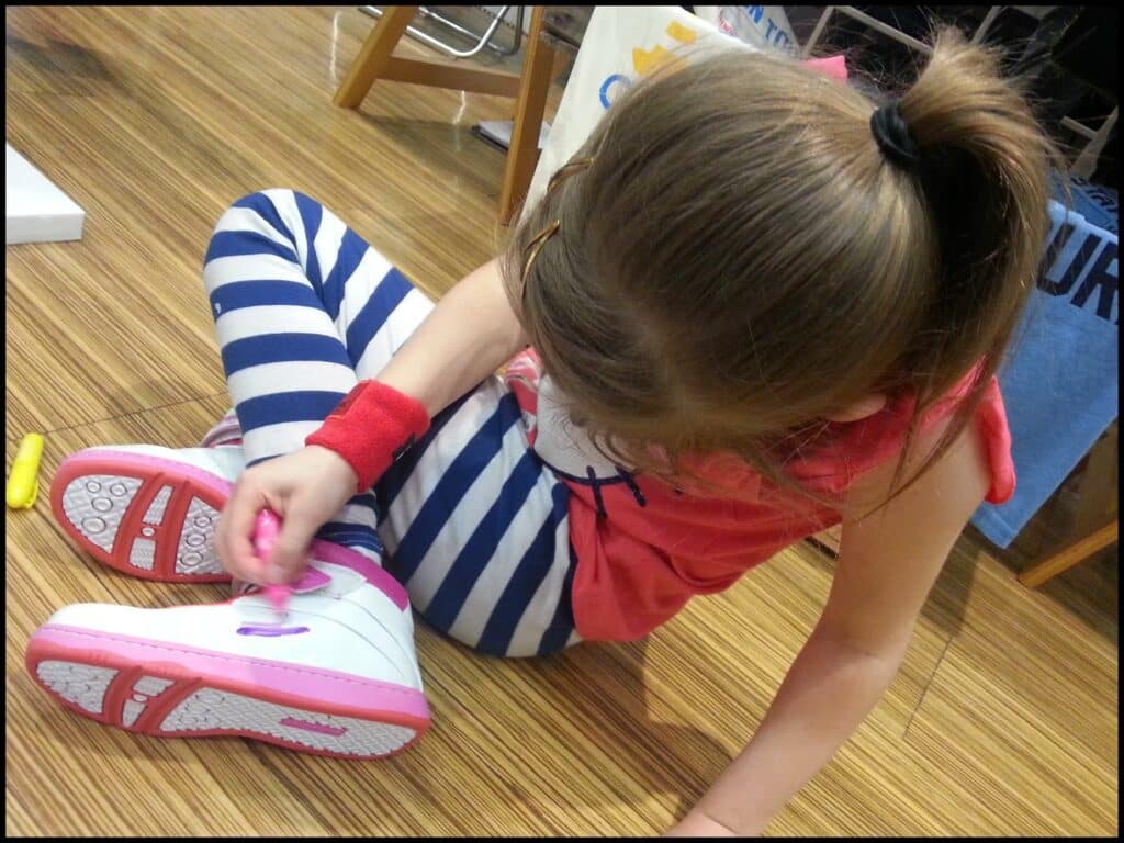 A young girl with light brown hair in a ponytail sits on a wooden floor, wearing a red shirt and blue-and-white striped leggings. She is colouring on her white trainer with a pink marker, focusing intently on her work.