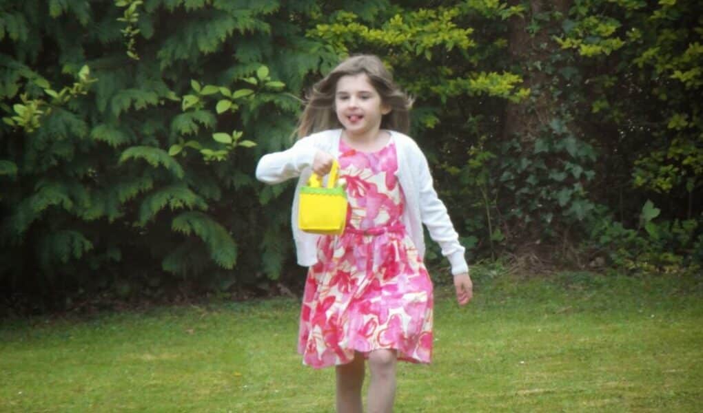 A young girl with long brown hair runs barefoot on grass. She is wearing a white cardigan over a pink and white floral dress and carries a small yellow handbag. Lush green bushes and trees fill the background.