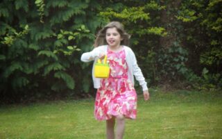 A young girl with long brown hair runs barefoot on grass. She is wearing a white cardigan over a pink and white floral dress and carries a small yellow handbag. Lush green bushes and trees fill the background.