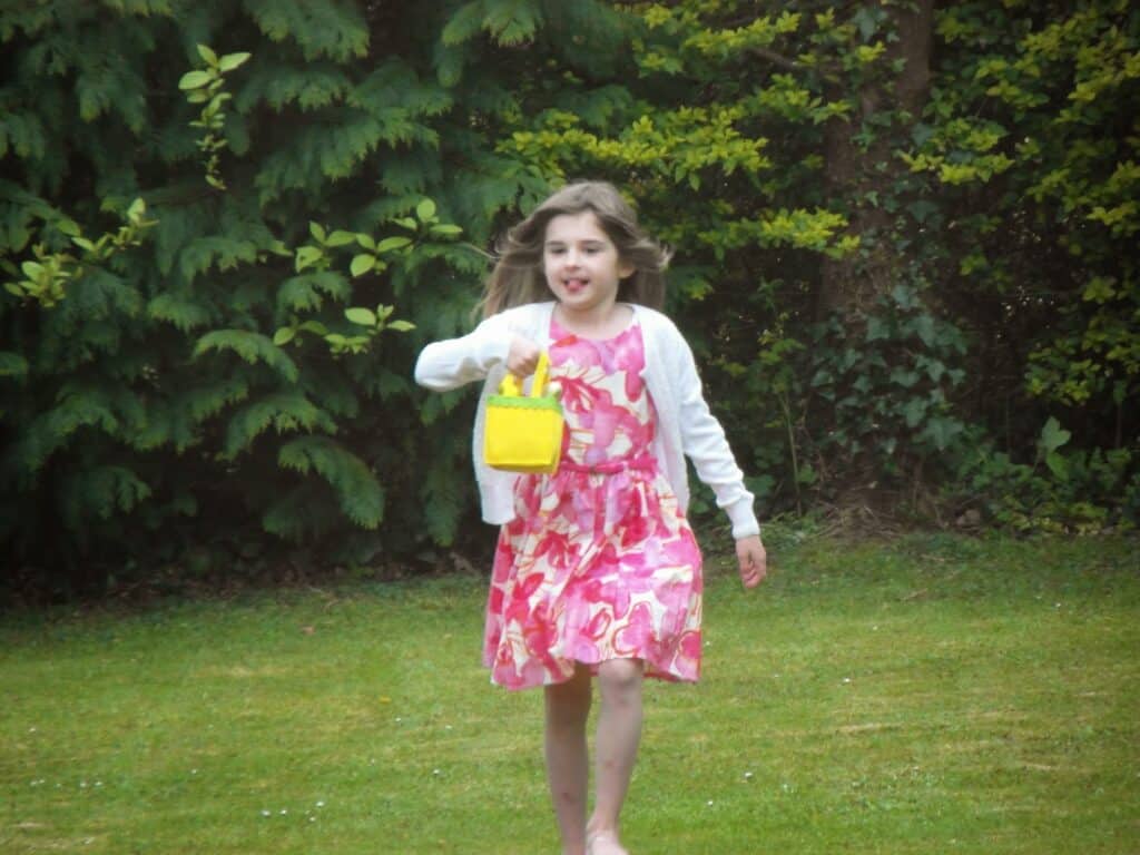 A young girl with long brown hair wears a white cardigan and a pink floral dress. She holds a small yellow handbag and walks barefoot on grass, surrounded by lush green bushes and trees. She has a joyful expression with her mouth slightly open.