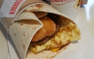 A McDonald’s breakfast wrap sits on a serviette. The wrap contains a folded omelette, melted cheese, a sausage patty, and a crispy hash brown inside a tortilla. Behind it, there is a large McDonald’s drink cup and a brown paper bag with logos.