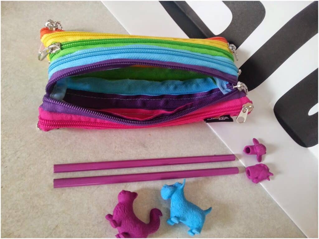 A colourful, rainbow-striped zippered pencil case sits open on a light surface. Below it are two purple pencils and three purple rubbers shaped like animals: a cat, a dog, and a pig. Part of a black-and-white object appears in the corner.