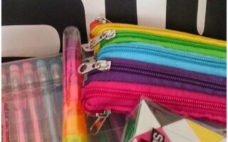 A close-up of colourful school supplies: three zip pouches in blue, green, and pink, a set of gel pens, a rainbow highlighter, and a black, white, and yellow snake puzzle. The background features bold black and white text.