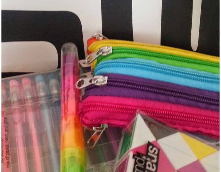 A close-up of colourful school supplies: three zip pouches in blue, green, and pink, a set of gel pens, a rainbow highlighter, and a black, white, and yellow snake puzzle. The background features bold black and white text.