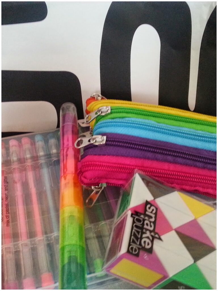 A close-up of colourful stationery items including a pack of gel pens, a multi-coloured pen, three zippered pencil cases (red, purple, and blue), and a geometric patterned snake puzzle, all set against a bold black and white background.