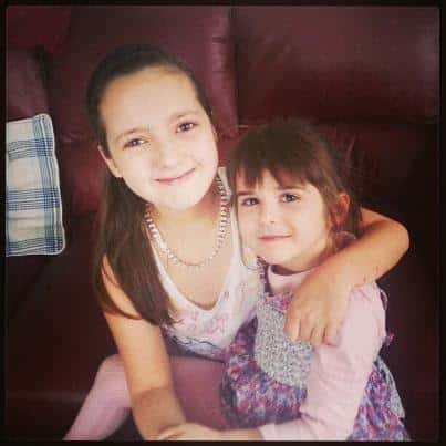 Two young girls sit closely together on a maroon sofa, smiling at the camera. The elder girl has long brown hair, wears a necklace, and hugs the younger girl, who has a fringe and wears a purple dress. A checked cushion is visible to the left.