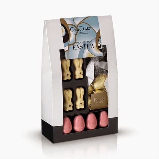 A gift box containing white chocolate bunny shapes, a gold-foiled bunny, and five pink foil-wrapped eggs. The packaging has a decorative label reading “Pick Me Up Easter” with pastel colours and a clear window showing the chocolates inside.