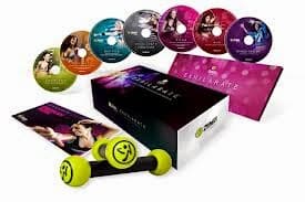 A Zumba fitness kit includes seven colourful workout DVDs, a programme guide, and a pair of black and lime green Zumba-branded dumbbells, all displayed around a branded box with a vibrant pink and purple design in the background.