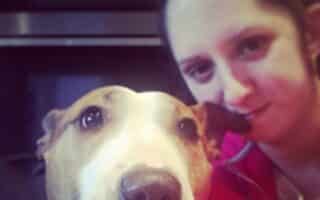 A woman with dark hair tied back smiles softly while taking a selfie with a brown and white Bull Terrier dog. The dog looks directly at the camera with a calm expression. The background is indoors, featuring a dark appliance or cupboard.
