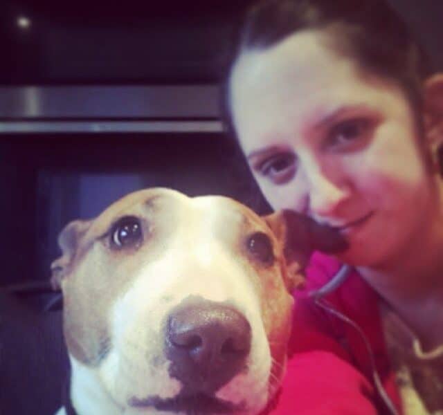 A woman with dark hair tied back smiles softly while taking a selfie with a brown and white Bull Terrier dog. The dog looks directly at the camera with a calm expression. The background is indoors, featuring a dark appliance or cupboard.