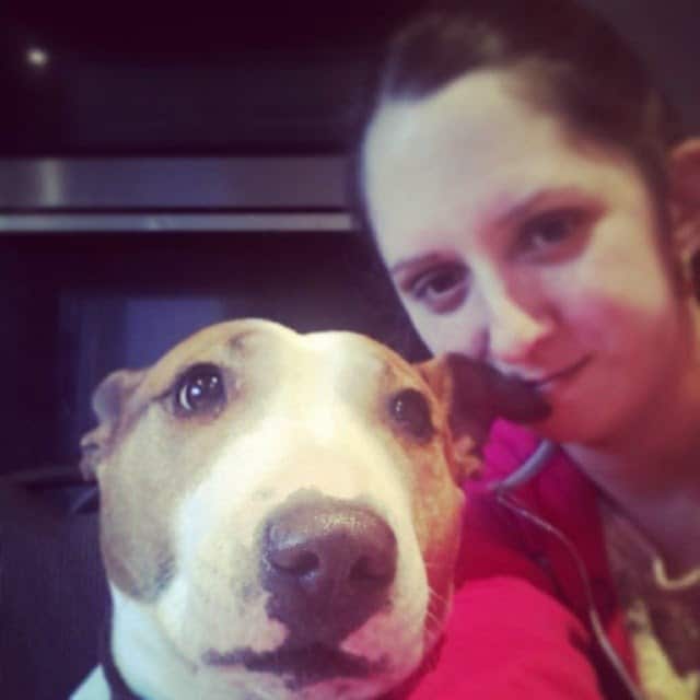 A woman with dark hair tied back smiles softly while taking a selfie with a brown and white Bull Terrier dog. The dog looks directly at the camera with a calm expression. The background is indoors, featuring a dark appliance or cupboard.