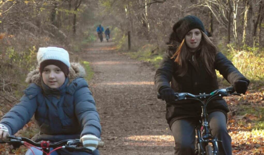 Two girls wearing coats, hats, and gloves ride bicycles on a leaf-covered forest path in autumn. Bare trees line the sunlit trail, and two distant figures are walking in the background. The scene feels cool and tranquil.