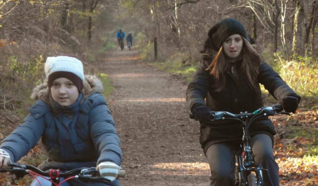 Two girls wearing coats, hats, and gloves ride bicycles on a leaf-covered forest path in autumn. Bare trees line the sunlit trail, and two distant figures are walking in the background. The scene feels cool and tranquil.