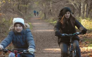Two girls wearing coats, hats, and gloves ride bicycles on a leaf-covered forest path in autumn. Bare trees line the sunlit trail, and two distant figures are walking in the background. The scene feels cool and tranquil.
