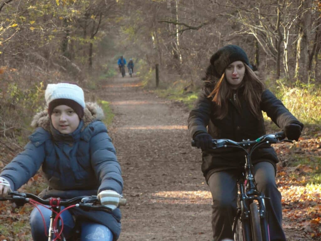 Two girls wearing winter coats, hats, and gloves ride bicycles on a forest path covered with dry leaves. Bare trees line the trail. In the background, three people walk together further down the path, surrounded by woodland in late autumn or early winter.