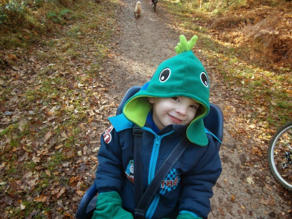 A young child in a blue dinosaur-themed hoodie sits in a child bike seat on a forest path. The child smiles at the camera. Fallen leaves cover the ground, and a bicycle wheel and a dog walking ahead are visible in the background.