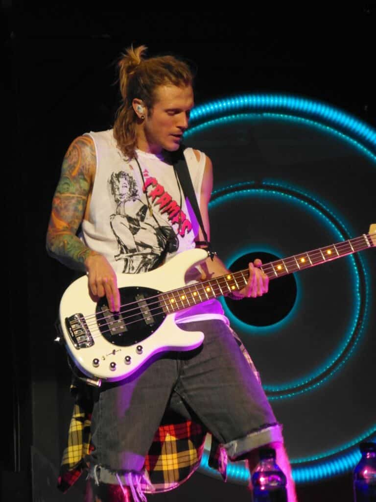 A musician with long, light brown hair in a half-up style plays a white electric bass guitar onstage. He wears a sleeveless shirt with a graphic print, ripped denim shorts, and has colourful tattoos on his arm. Neon blue rings glow in the dark background.
