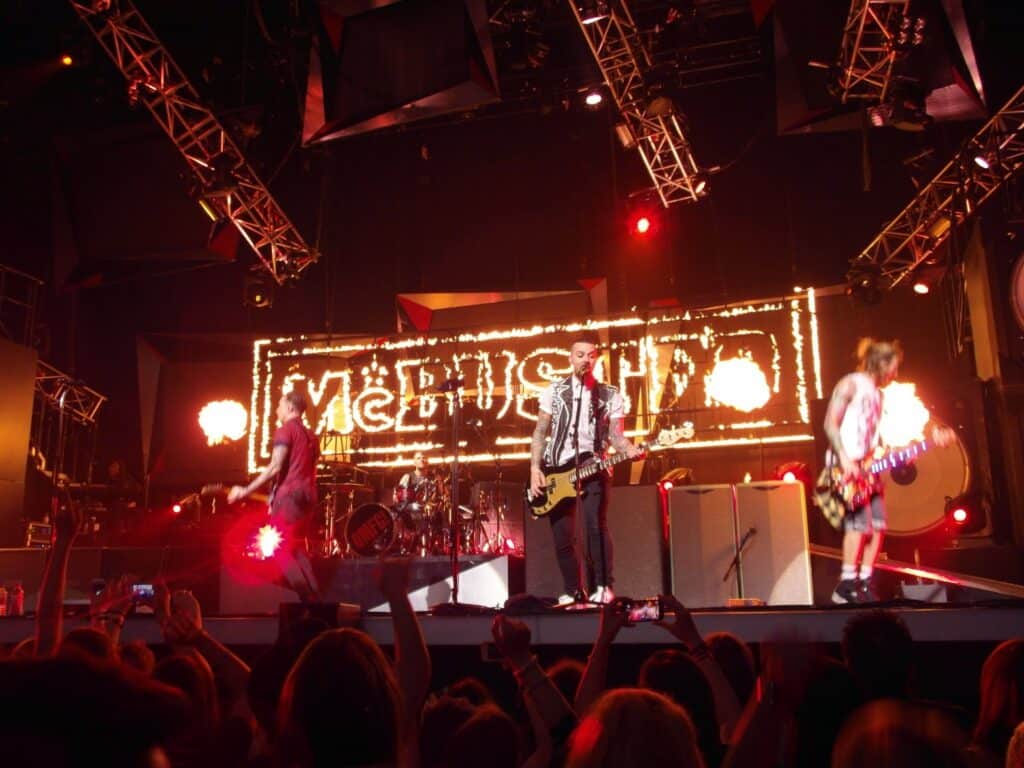 A rock band performs on stage under bright red lights. Three guitarists are visible, each playing their instruments energetically. Behind them, a large illuminated sign reads "MCBUSTED," and an enthusiastic crowd is visible in the foreground.