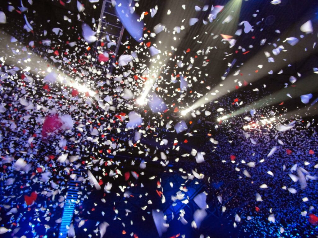 Colourful confetti, mostly white with some red pieces, fills the air under bright stage lights at a concert or celebration. The background is dark with hints of blue lighting, and the confetti appears to be drifting down from above.