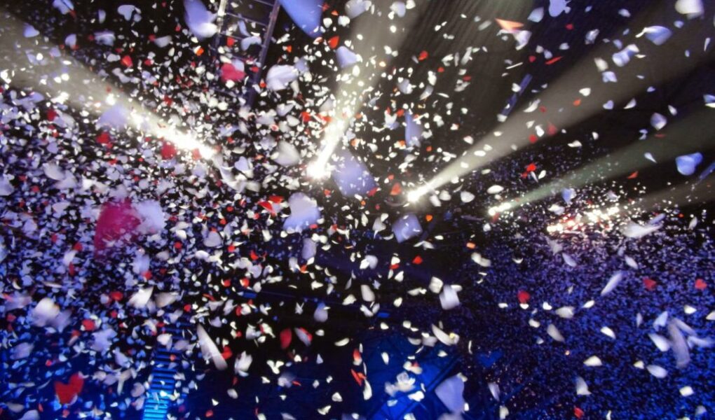 Hundreds of white, red, and pink confetti pieces float through the air under bright stage lights, creating a festive atmosphere above a dark, crowded concert or event venue. Blue lighting highlights the audience below the swirling confetti.