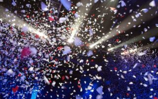 Hundreds of white, red, and pink confetti pieces float through the air under bright stage lights, creating a festive atmosphere above a dark, crowded concert or event venue. Blue lighting highlights the audience below the swirling confetti.
