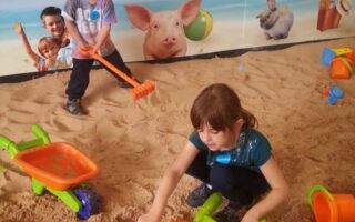 Two young children play with colourful wheelbarrows and sand toys in an indoor sandpit. A girl scoops sand, and a boy holds a sand rake. The backdrop shows animals and reads “Umberslade Beach Party.” Toys and buckets are scattered in the sand.