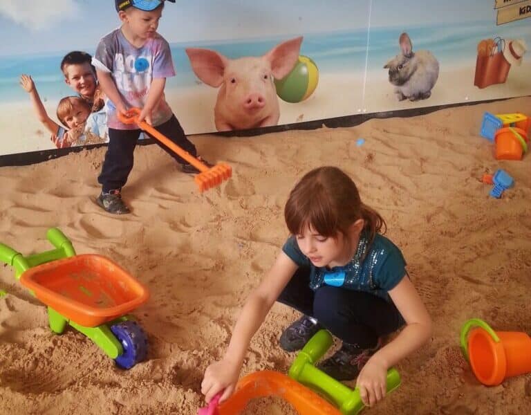 Two young children play with colourful wheelbarrows and sand toys in an indoor sandpit. A girl scoops sand, and a boy holds a sand rake. The backdrop shows animals and reads “Umberslade Beach Party.” Toys and buckets are scattered in the sand.