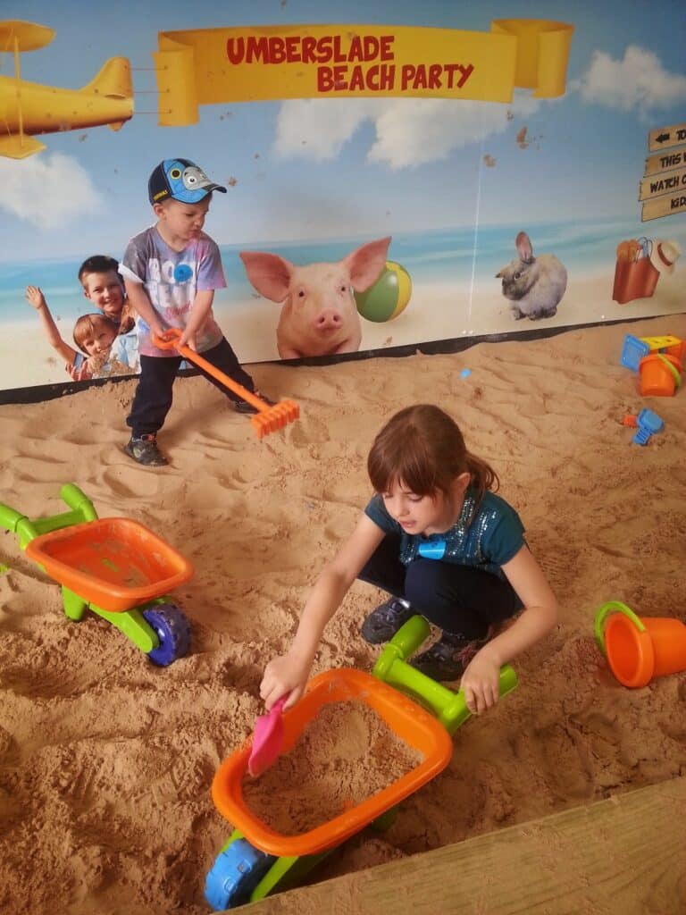 Two children play in an indoor sandpit with toy wheelbarrows, spades, and buckets. A mural behind them shows smiling children, a pig, and a rabbit on a beach, with a banner reading "Umberslade Beach Party." Sand toys are scattered around.