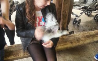 A young girl with long brown hair, wearing a black jacket, black leggings, and bright pink trainers, sits on a wooden bench indoors. She smiles and holds a small, white-feathered animal. Blurred people and a pushchair are visible in the background.