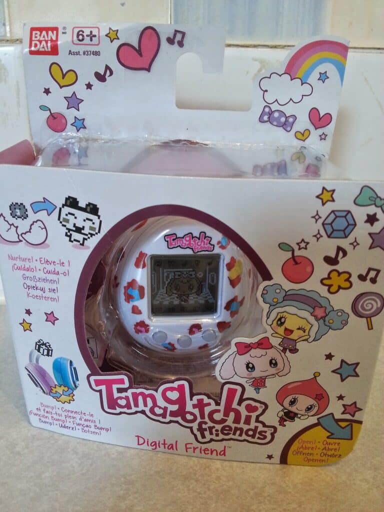 A Bandai Tamagotchi Friends digital pet in original packaging. The white egg-shaped device with colourful hearts is visible through a window. The box features cute cartoon characters, hearts, stars, and colourful doodles, with text in several languages.