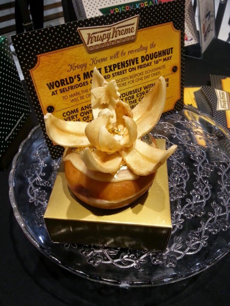 A large glazed doughnut sits on a glass plate with a golden flower decoration on top. A sign behind it reads, “Krispy Kreme: World’s Most Expensive Doughnut,” describing its debut at Selfridges, London. The set-up looks elegant and luxurious.