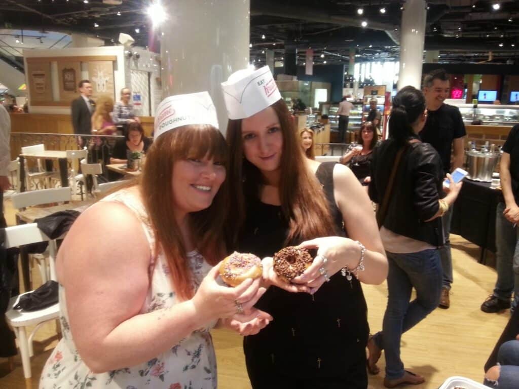 Two women wearing Krispy Kreme paper hats smile and hold decorated doughnuts towards the camera in a busy, modern café. Other people are seated and standing in the background, and the atmosphere is bright and lively.