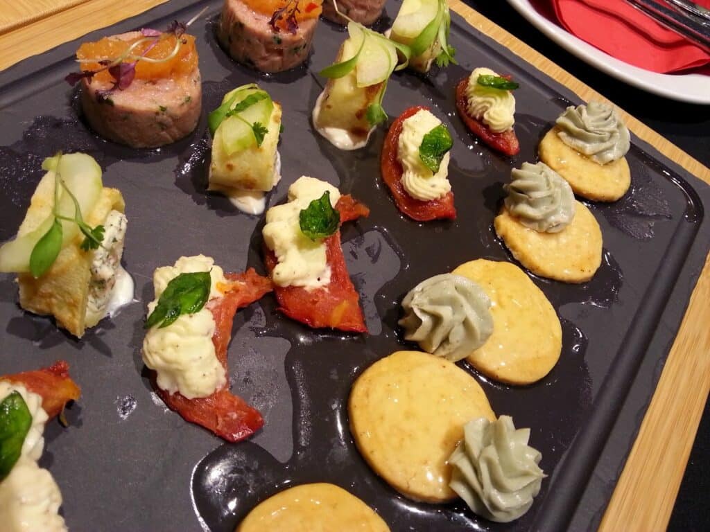 A black platter displays assorted gourmet appetisers: round meat medallions with herbs, bites topped with cucumber and apple slices, crackers with piped green and white mousses, and roasted tomato pieces garnished with microgreens, set on a wooden board.