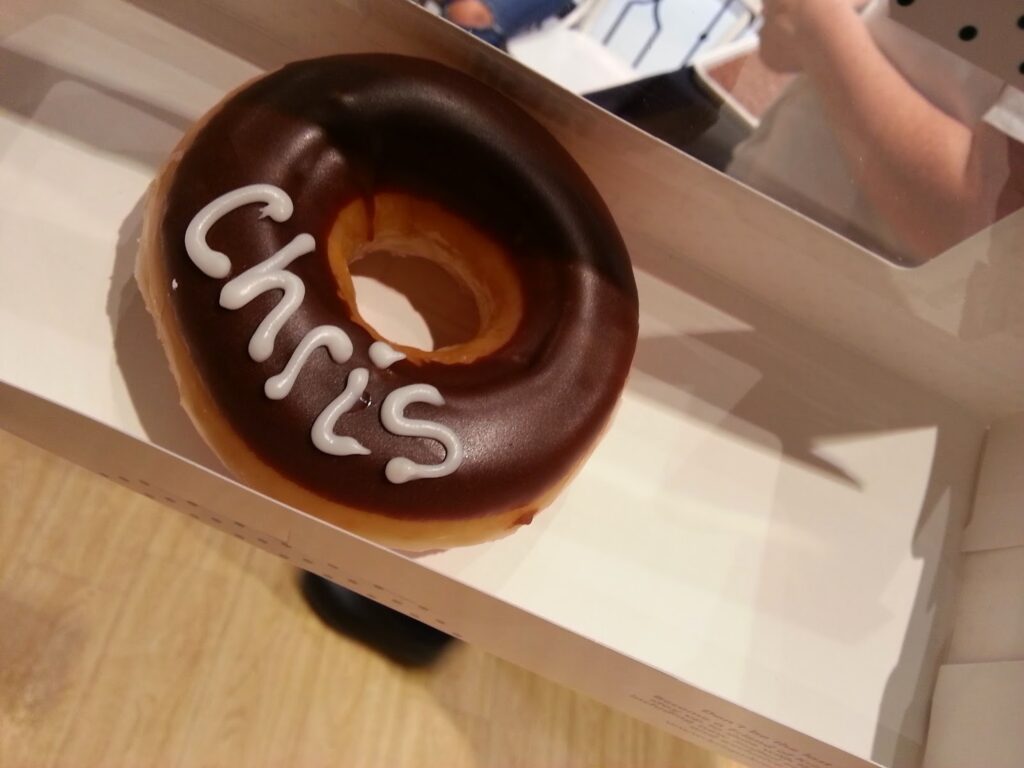 A chocolate-covered doughnut with the name "Chris" written in white icing sits inside a white box on a wooden surface. Part of a person's arm and a phone are visible in the background, reflected in the shiny surface of the box lid.