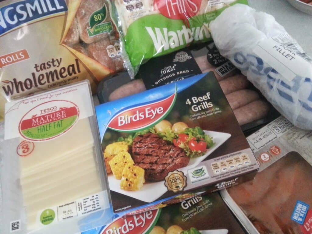 An assortment of packaged foods including Birds Eye 4 Beef Grillsteaks, Tesco Mature Half Fat cheese slices, Warburtons bread rolls, sausages, fish fillet, and Kingsmill wholemeal rolls, all displayed together on a surface.