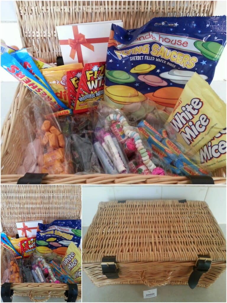 A wicker hamper filled with assorted retro sweets in colourful wrappers, including Flying Saucers, Fizz Wiz, White Mice, candy necklaces, and chewy bars. The lid has black leather straps and a card with a red ribbon is inside.