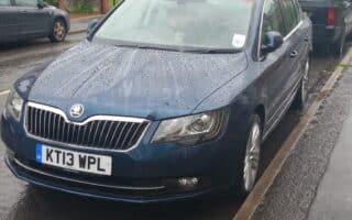 A dark blue Skoda car with UK number plate KT13 WPL is parked on a wet street beside a pavement. Rain droplets are visible on the car and road. Other cars and brick blocks of flats with trees are in the background.