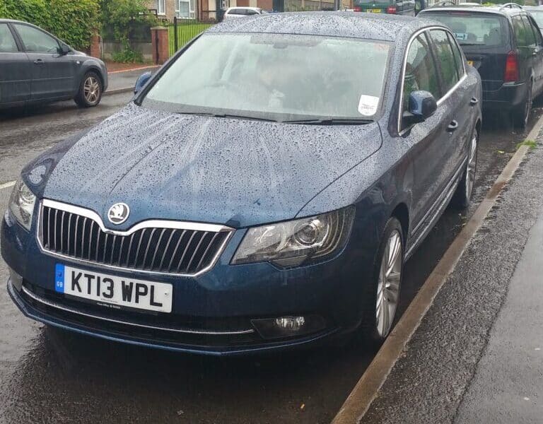 A dark blue Skoda car with UK number plate KT13 WPL is parked on a wet street beside a pavement. Rain droplets are visible on the car and road. Other cars and brick blocks of flats with trees are in the background.