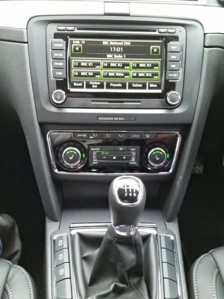 Car dashboard interior showing a digital radio display tuned to BBC National DAB, dual climate controls with dials for temperature and fan speed, buttons for air conditioning, and a manual gear lever with a 6-speed pattern. Black leather trim surrounds the controls.