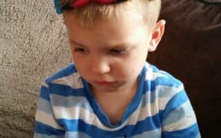 A young boy with short blond hair, wearing a blue and white striped shirt, sits on a sofa looking down with a serious expression. He has a colourful homemade crown of pipe cleaners and googly eyes on his head.