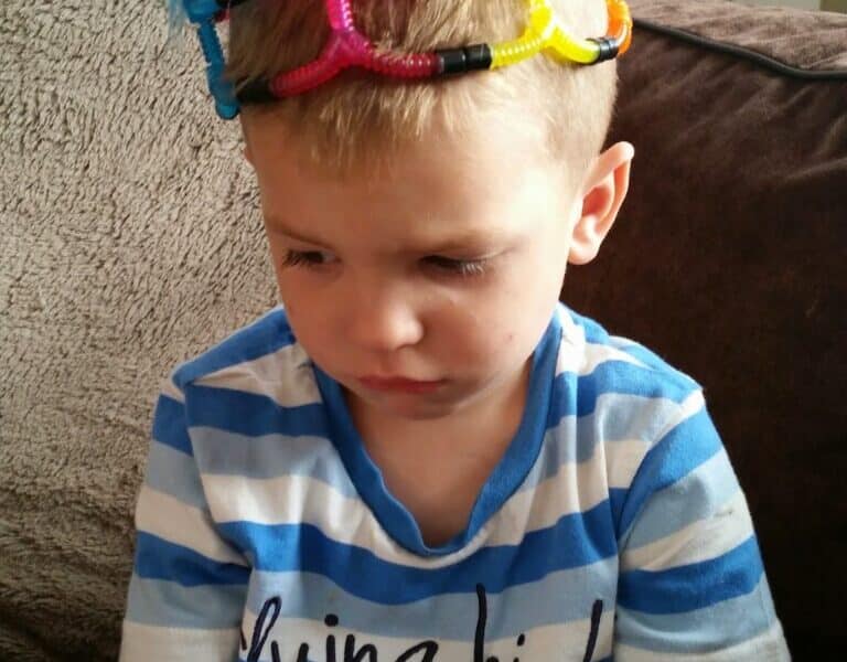 A young boy with short blond hair, wearing a blue and white striped shirt, sits on a sofa looking down with a serious expression. He has a colourful homemade crown of pipe cleaners and googly eyes on his head.