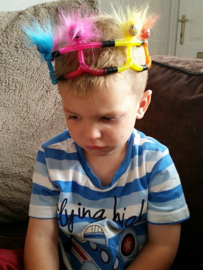 A young boy with blond hair, wearing a blue and white striped shirt with an aeroplane graphic, sits on a sofa looking down with a sad expression. He has a colourful, playful crown made of pipe cleaners and googly eyes on his head.