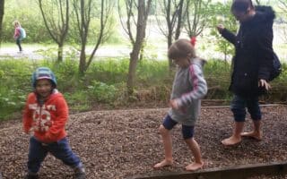 Four children and a woman walk barefoot on a gravel path outdoors. The children wear jackets and one wears a red hood. Trees and greenery surround them, and another child and adult walk in the background. All seem to be enjoying the activity.
