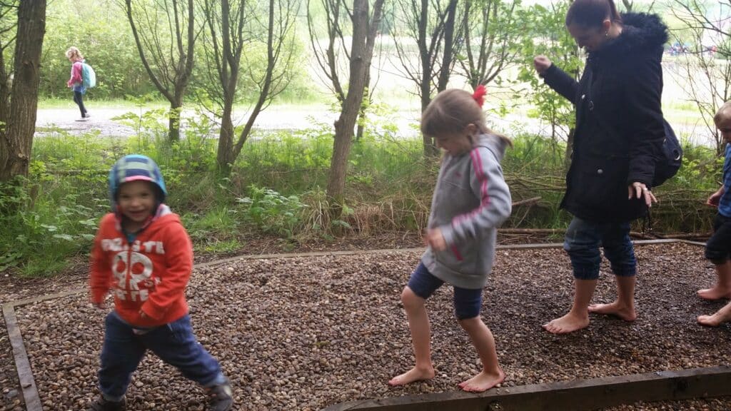 Four children and a woman walk barefoot on a gravel path outdoors. The children wear jackets and one wears a red hood. Trees and greenery surround them, and another child and adult walk in the background. All seem to be enjoying the activity.