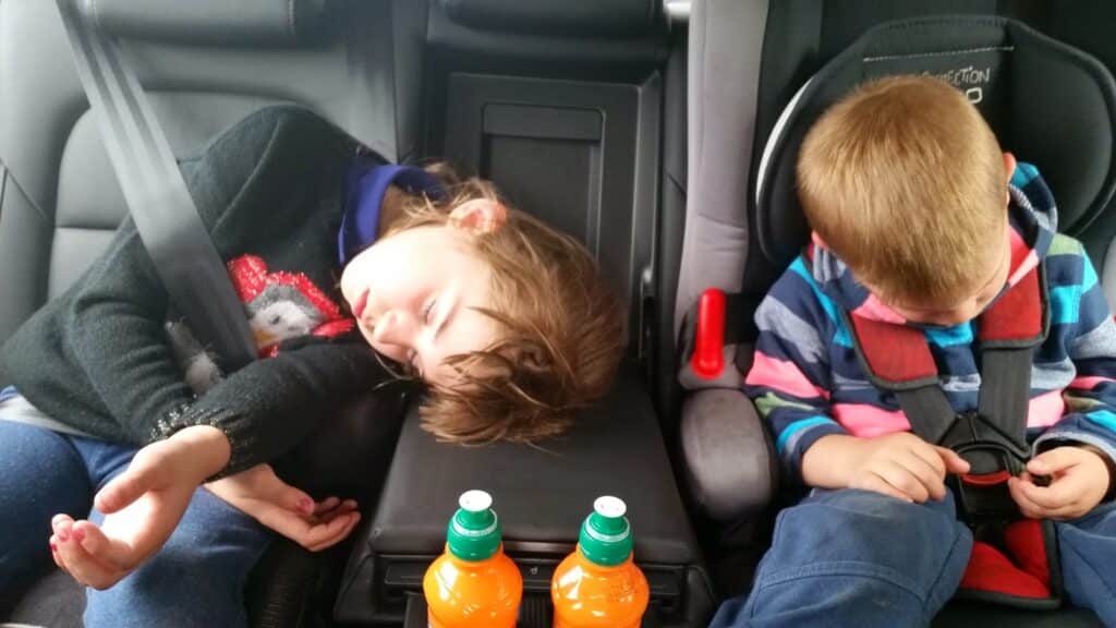 Two young children in the back seat of a car, both asleep. The girl on the left leans her head on the centre armrest, and the boy on the right, in a striped hoodie and car seat, tilts his head down. Two orange drink bottles sit in front of them.