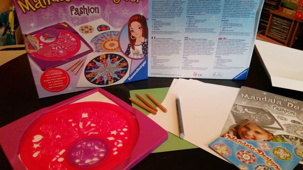 A Mandala designer kit is spread out on a table, showing a pink stencil, brown coloured pencils, white paper, an instruction booklet, and the product packaging with colourful mandala designs and an illustrated girl on the front.