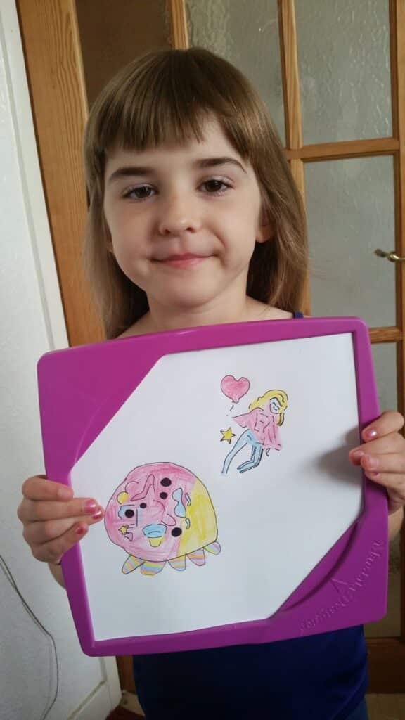 A young girl with straight light brown hair and fringe smiles slightly while holding up a white drawing board with a pink border. The board displays two colourful, childlike drawings: a round character and a blonde girl with a pink heart.